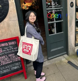 A woman holding a tote bag with a red logo in front of a bookshop's door.