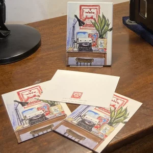 A stack of copies of the same postcard picturing the shop's typewriter on its desk, surrounded by a plant, a lamp, and the logo of the shop.